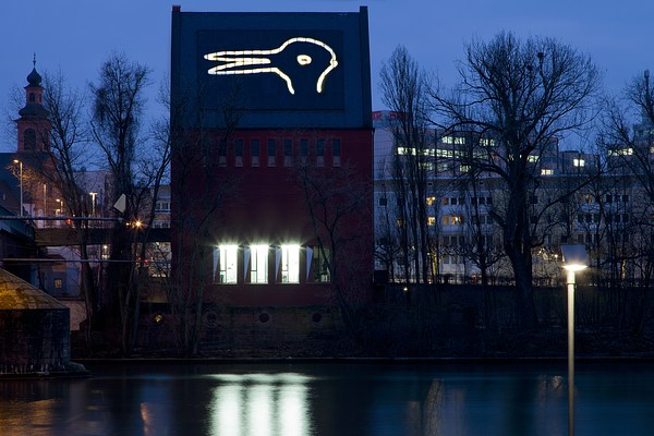 Exterior view of the tall, red building at night, the white line drawing on its roof illuminated. The grey roof is all dark, but the line drawing is glowing. The Duck/Bunny image reads more clearly and stands out from the surrounding neighborhood and skyline.