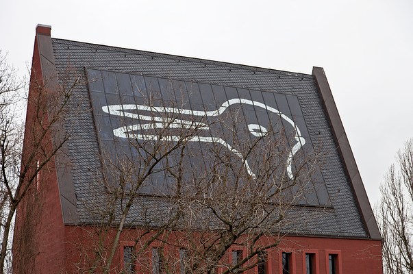 A closer view of the large white line drawing titled "Duck/Bunny" on the roof of the tall red building. If you look at the drawing one way it appears to be a duck, but the beak of the duck can also appear as the ears of a bunny if it's looked at another way. The sky around the building is pale gray and the trees are all bare of leaves.