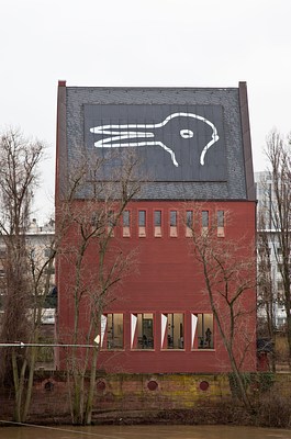 Exterior view of a tall red building with a peaked grey roof. On the grey roof is a huge white line drawing titled, "Duck/Bunny." If you look at the drawing one way it appears to be a duck, but the beak of the duck can also appear as the ears of a bunny if it's looked at another way. The sky around the building is pale gray and the trees are all bare of leaves.