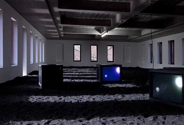 Installation view of a dimly lit gallery. The walls are white with numerous vertical inset windows and three large, cube-shaped monitors on a floor covered with sparkling black sand. The monitors show two brightly illuminated spheres, one larger and one smaller, that appear planetary or celestial.