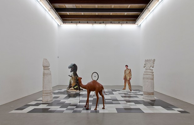 Installation view of a large white gallery space with a gray concrete floor and florescent lights along the perimeter of the ceiling. On the floor is a large square made up of an irregular pattern of smaller squares in different shades of white, ivory, black, and gray. Large cutouts are arranged on the board like game pieces, making the installation seem like a life-size chess board. The cutouts are of a camel, a dinosaur, Elvis in a gold suit, a replica of a sculpture by Duchamp of a white stool with an inverted wheel mounted on top of it, and two white shapes that appear more like traditional chess pieces, but enlarged.