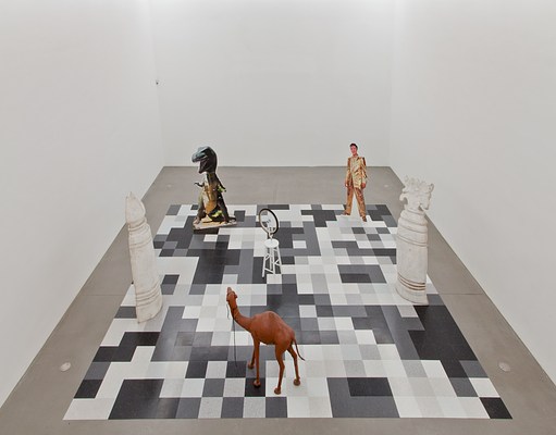 Another overhead view of the chessboard-like installation with the whole board visible. In the center of the board is the replica of Duchamp's wheel sculpture. Surrounding it at the 6 o'clock position is the camel sculpture. At 9 o'clock is one of the enlarged, white chess pieces. At 11 o'clock is the tyrannosaurus rex cutout. At 1 o'clock is the cutout of Elvis in a gold suit. And at 3 o'clock is the other chess piece.
