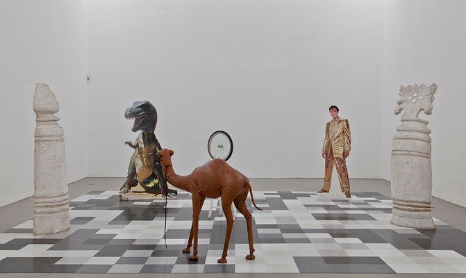 Another view of the chessboard-like installation taken from eye level. Directly in front of us is the camel, whose body partially obscures the white stool behind it, the bicycle wheel appearing to float out of the its hump. To the left and right are two enlarged, white chess pieces. The t-rex and Elvis cutouts stand behind.