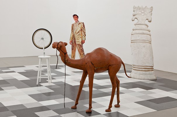 Another view of the chessboard-like installation taken from a position within the board. We see the camel in the foreground, its neck angled toward the wheel sculpture. Elvis and one of the enlarged, white chess pieces stand behind.