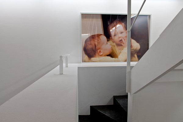 A large photograph with a white frame hangs in a white stairwell at a place where the steps begin to wind. The photo shows a toddler with light skin and red hair standing with their hands pressed against a mirrored surface. On the left side of the image, the toddler's head is shown in profile, looking directly at the mirror. On the right side of the image, we see the toddler's reflection looking directly out at us.