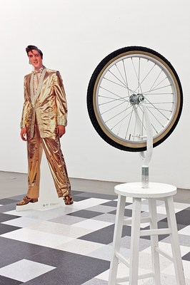 Another view of the chessboard-like installation showing a close up of the replica of Duchamp's wheel sculpture in the foreground and the cutout of Elvis in a gold suit in the background. On closer inspection, the smaller squares that make up the larger game board appear to be linoleum tiles commonly used in kitchens or other public or commercial spaces.