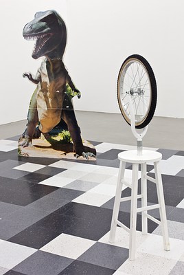 Another view of the chessboard-like installation showing a close up of Duchamp's wheel sculpture in the foreground and the green t-rex cutout in the background. The t-rex doesn't look very realistic. Rather, it looks like a replica one might see in a diorama at a natural history museum, or at a theme park.