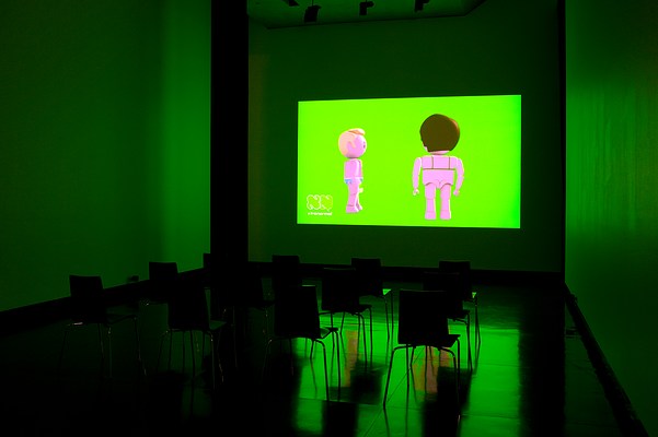 Installation view of the same dark room with three walls, ceiling and floor visible as in previous image but seen from the right. On the center wall we can see a large video projection. In the video still, we can see two animated figures in full body against a green screen. The figures are doll-like, similar to those in Playmobil or LEGO. The figure to the left is male. He is seen in profile, turned to the right. He has light skin, short blonde quiff, and is wearing baby blue briefs with a white lining. To the left of him, slightly closer to us, we can see what appears as a female figure. She is facing away from us, and we can see green bra straps on her back. She has light skin, and short brown hair. In the left bottom corner of the projection, we can see a logo that looks like the outline of two capital letters "N" in white cartoonish font. Underneath it, we can read the text "xtranormal" in white modern font. In front of the projection, four black chairs with square backrests and chrome legs stand in rows of three. To the left, obscuring the corner where the center wall and the left wall meet, we can see a narrow black curtain stretching from just below the ceiling to floor. The floor is dark and polished, and reflects the green light from the projection. In the ceiling, we can see spot lights that are turned off.
