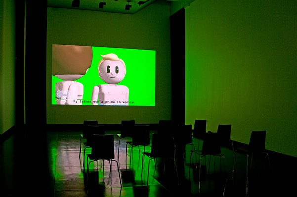 Installation view of the same dark room with three walls, ceiling and floor visible as in previous image but seen from the right. On the center wall we can see a large video projection. In the video still, we can see two animated figures from the waist up against a green screen. The figures are doll-like, similar to those in Playmobil or LEGO. To the left in the projection, we can see a female figure. Her torso is seen in profile, while her head is turned to the left. Her skin is light and her brown hair is cut in a short bob. We can't se her face, as she is looking at a male figure slightly further away from us, in the center of the projection. He has light skin, short blonde quiff, and is looking straight at us. His upper body is bare and we can see his nipples. His head is round and his eyes are two vertical oval black shapes with white triangles, representing reflecting light. He has fine light brown eye lashes. His mouth is a thin, vertical black line and his nose is in the shape of a small button. There is no particular facial expression to be discerned. In the left bottom corner of the projection, we can see a logo that looks like the outline of two capital letters "N" in white cartoonish font. Underneath it, we can read the text "xtranormal" in white modern font. At the bottom of the projection, we can see the captions "My father won a price in Venice." in dark grey typewriter font. In front of the projection, four black chairs with square backrests and chrome legs stand in rows of three. On each side of the projection in the corners where the center wall meets the left and the right wall, we can see narrow black curtains stretching from just below the ceiling to floor. The floor is dark and polished, and reflects the green light from the projection. In the ceiling, we can see spot lights that are turned off.