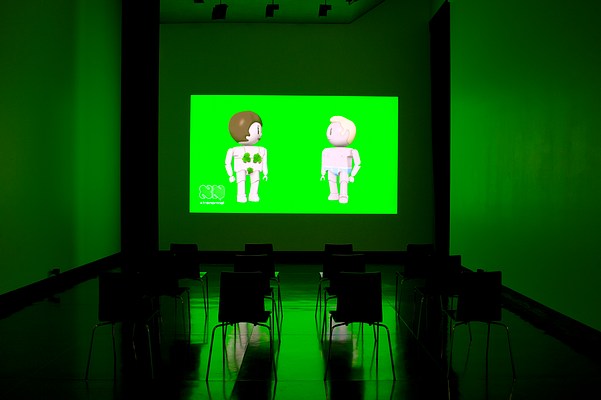 Installation view of the same dark room with three walls, ceiling and floor visible as in previous image but seen from straight ahead. On the center wall we can see a large video projection. In the video still, two animated figures in full body against a green screen. The figures are doll-like, similar to those in Playmobil or LEGO. The figure to the left is male. He is seen in profile, turned to the right. He has light skin, a short blonde quiff, and is wearing baby blue briefs with a white lining. To the left of him, slightly closer to us, we can see what appears as a female figure. She is facing away from us, and is wearing a green bra. She has light skin, and short brown hair. In the left bottom corner of the projection, we can see a logo that looks like the outline of two capital letters "N" in white cartoonish font. Underneath it, we can read the text "xtranormal" in white modern font. At the bottom of the projection, we can see the captions "The more you fuck, the better you will edit" in grey typewriter font. In front of the projection, four black chairs with square backrests and chrome legs stand in rows of three. On each side of the projection in the corners where the center wall meets the left and the right wall, we can see narrow black curtains stretching from just below the ceiling to floor. The floor is dark and polished, and reflects the green light from the projection. In the ceiling, we can see spot lights that are turned off.