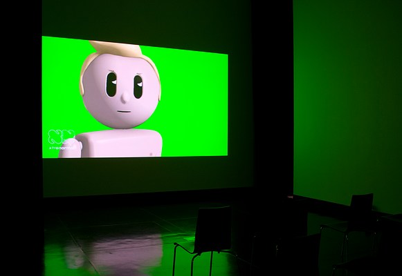 Installation view of the same dark room with three walls, ceiling and floor visible as in previous image but seen from the left. On the center wall we can see a large video projection. In the video still, we can see a male animated figure from just below the shoulders up against a green screen. He is positioned slightly to the right in the frame. The figure is doll-like, similar to those in Playmobil or LEGO. His head is round, and he has a blonde quiff which is slightly out of frame and cropped. His eyes are two vertical black ovals with white triangles, representing reflecting light. He has fine light brown eye lashes. His mouth is a thin, vertical black line and his nose is in the shape of a small button. There is no particular facial expression to be discerned. In the left bottom corner of the projection, we can see a logo that looks like the outline of two capital letters "N" in white cartoonish font. Underneath it, we can read the text "xtranormal" in white modern font. In front of the projection, we can see three black chairs with square backrests and chrome legs in a row. On each side of the projection in the corners where the center wall meets the left and the right wall, we can see narrow black curtains stretching from just below the ceiling to floor. The floor is dark and polished, and reflects the green light from the projection.