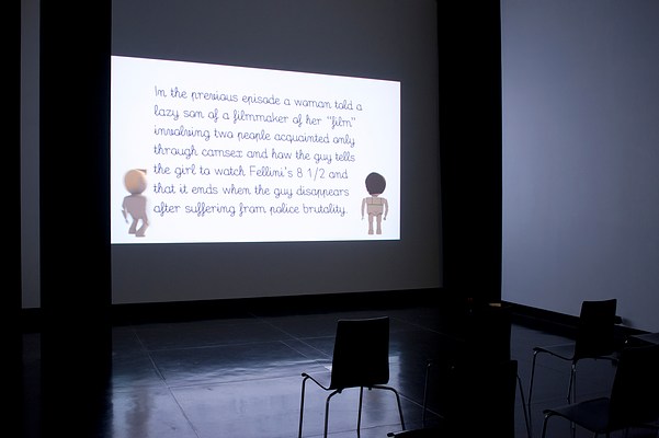 Installation view of the same dark room but seen from the right. On the center wall we can see a large video projection. In the video still, we can see a text passage against a white background and two animated figures standing in each corner, framing the text. The figures are doll-like, similar to those in Playmobil or LEGO. In the bottom right corner of the projection, we can see a female figure in full body. Her skin is light and her brown hair is cut in a short bob. She is turned away from us. On her back, we can see thin green bra straps. In the bottom right corner of the projection, we can see a male figure. He has light skin, a short blonde quiff, and is seen from behind but turned slightly to his right. His figure is blurry, suggesting movement, as if he is taking a step forward. The text passage is laid out in seven justified rows. The font is black and appears to be mimicking handwriting. The passage reads: "In the previous episode a woman told a lazy son of a filmmaker of her "film" involving two people acquainted only through camsex and how the guy tells the girl to watch Fellini's 8 1/2 and that it ends when the guy disappears after suffering from police brutality." In front of the projection, three black chairs with square backrests and chrome legs stand in a row. On each side of the projection in the corners where the center wall meets the left and the right wall, we can see narrow black curtains stretching from just below the ceiling to floor. The floor is dark and polished, and reflects the white light from the projection.