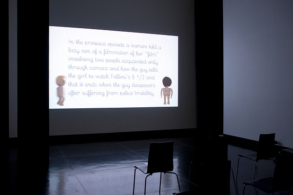 Installation view of the same dark room but seen from the right. On the center wall we can see a large video projection. In the video still, we can see a text passage against a white background and two animated figures standing in each corner, framing the text. The figures are doll-like, similar to those in Playmobil or LEGO. In the bottom right corner of the projection, we can see a female figure in full body. Her skin is light and her brown hair is cut in a short bob. She is turned away from us. On her back, we can see thin green bra straps. In the bottom right corner of the projection, we can see a male figure. He has light skin, a short blonde quiff, and is seen from behind but turned slightly to his right. His figure is blurry, suggesting movement, as if he is taking a step forward. The text passage is laid out in seven justified rows. The font is black and appears to be mimicking handwriting. The passage reads: "In the previous episode a woman told a lazy son of a filmmaker of her "film" involving two people acquainted only through camsex and how the guy tells the girl to watch Fellini's 8 1/2 and that it ends when the guy disappears after suffering from police brutality." In front of the projection, three black chairs with square backrests and chrome legs stand in a row. On each side of the projection in the corners where the center wall meets the left and the right wall, we can see narrow black curtains stretching from just below the ceiling to floor. The floor is dark and polished, and reflects the white light from the projection.