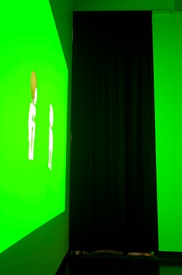 Close up of one of the rectangular black curtains seen in previous installation views. We can see two walls meeting in a corner in the center of the image. On the left wall, we can see a video projection with two animated figures in full body against a green screen. The figures are doll-like, similar to those in Playmobil or LEGO. They are standing next to each other, facing each other. On the right wall, a black narrow curtain hangs positioned with its left edge in the corner. To the right, we can see some of the right wall reflecting the green light from the projection. Underneath the curtain, we can glimpse some warm light, suggesting that the curtain might be hiding a doorway to another room.