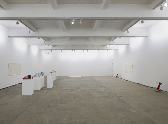 Installation view of a large, open gallery space with white walls and ceiling beams, and a beige concrete floor. On our left is a cluster of white pedestals and an off-white rectangular artwork hanging on the wall behind them. Directly in front of us, there appears to be another off-white rectangular piece lying on the floor in the distance. And on our right is a third off-white rectangular piece hanging on the wall, and two portable red space heaters on the ground pointing towards the center of the gallery.