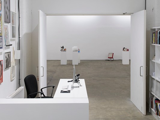 View from inside an office through a doorway into the gallery, where there are four white pedestals and a portable red space heater on the floor. The pedestals seem to have an assortment of small items heaped together on top of them, but they are too far away to see in any detail. In the office, framed pictures and a tote bag hang on the wall behind a black office chair and desk, opposite which a white bookshelf is partially visible.