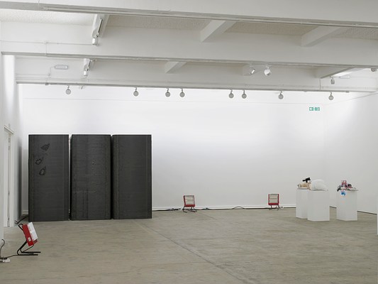 Installation view showing a row of three large, charcoal-colored foam blocks, the approximate height and width of a doorway, along with a cluster of three pedestals with an assortment of objects heaped on top of them, and three portable red space heaters, all pointing in towards the center of the gallery.