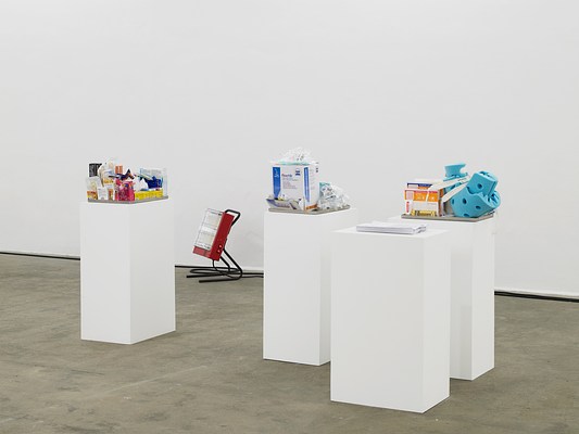 A closer view of the cluster of four pedestals with various items heaped on top of them. On closer inspection, the items look to be medical supplies and drugstore items like topical creams, bandages, and rubber gloves, all contained within shallow metal trays. One tray contains a large foam object that stands out because of its bright blue color. One of the four pedestals has a stack of papers on it. There is a portable red space heater in the background.