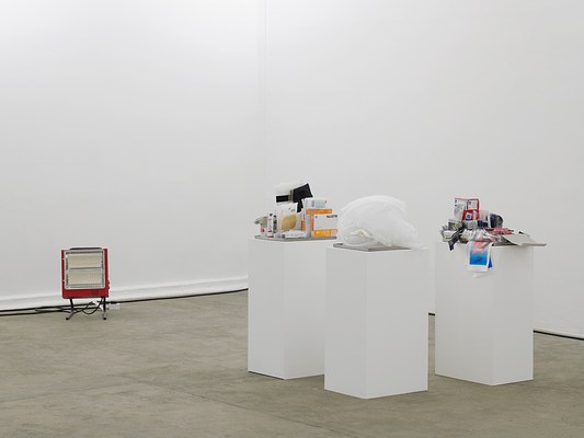 Closer view of three more pedestals with shallow metal trays containing various medical and drugstore items. On one there is a pile of what look like clear, plastic ventilator tubes. A portable red space heater sits on the floor to our left.
