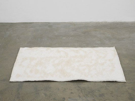 Close-up view of off-white rectangular artwork installed on the ground. It looks like it could be a thin sheet of foam or a thick sheet of paper, and it has a slight wave like paper that has gotten wet and then dried. It is much larger than a regular sheet of printer paper—more like the size of a welcome mat—and is mostly white with peachy-beige splotches. Its edges are rough and unfinished.