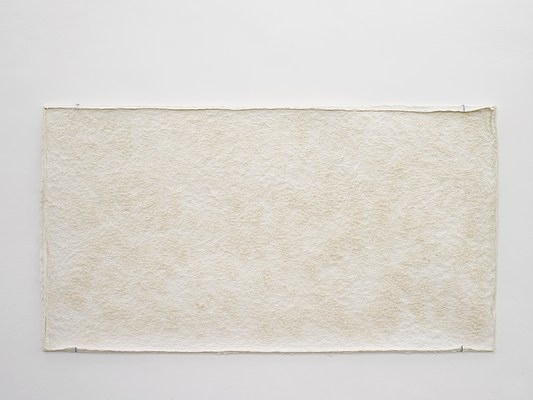 Close-up view of off-white rectangular wall-mounted work with bumpy texture and peachy-beige splotches against a white background. It looks like it could be made of some kind of foam or paper. Its edges are unfinished and pulpy.
