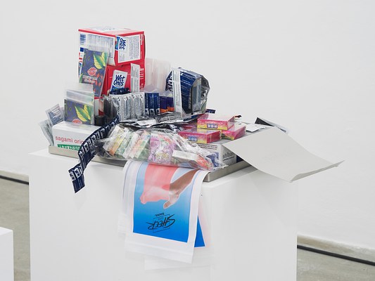 Close-up view of shallow metal tray containing various drugstore items, including packets of Wet Ones wipes, condoms, and Sheer Glyde Dams, on a white pedestal.