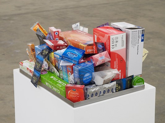 Close-up view of shallow metal tray with various drugstore items, including durex condoms, clingfilm, Vaseline, and rubber gloves, on a white pedestal.