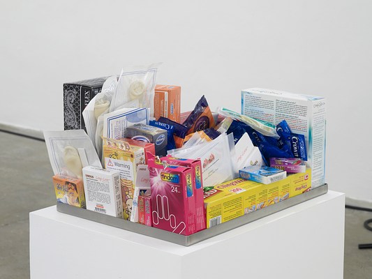 Close-up view of shallow metal tray with various drugstore sexual wellness items, including condoms, dams, and an at-home HIV test, on a white pedestal.