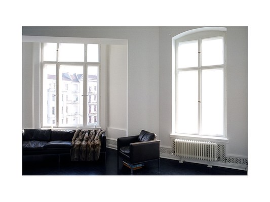 Installation view of a room with two large windows, white walls and black floor. We can see two walls meeting, forming a wide corner to the right in the picture. In the left half of the image, though a large square opening in the left wall, we can see the wall and a large window in another room. The window has six window panes in three vertical rows, the lower ones being about third as high as the upper. Through the window we can see the facades, windows and roofs of opposing buildings typical to a European city. The sky is light grey and the light is bright white. In front of the window stands a low black leather couch with three seats facing us. Over the seat to the right, a brown fur blanket or coat has been draped. The leather is reflecting the light from the window behind. To the right in the room, we can see a white wall with a rectangular window with four window panes in two vertical rows. Beneath the window hangs a white rectangular radiator. Along the base moulding of the room runs a grid with a pattern of small cross-shaped perforations. It continues behind the radiator and into the second room in the far right. Most likely it serves as a ventilation system. From the window emanates a white light. On the floor in front of the window, forming a group with the couch and facing to the left, stands an armchair in black leather. It matches the couch and appears to be designer furniture. Underneath the armchair, we can see a stack of either books or board games.