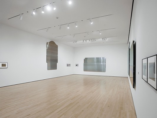 Installation view of an open gallery space with light blonde wood floors, white walls, and track lighting. The view is from the front right corner to the back left corner. Small, framed pictures too distant to discern are interspersed between large, silver-metallic wall works shaped like highway signs or other public markers. The signs are blank, bearing no text or imagery.