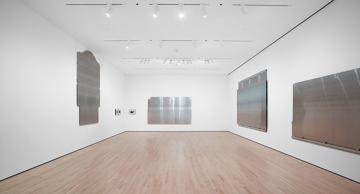 Installation view facing the back wall of the gallery. On our left is a vertically-oriented, silver-metallic wall work consisting of three, horizontal rectangular panels stacked on top of one another. The remaining three silver-metallic wall works are horizontally-oriented rectangles. One hangs on the wall directly in front of us, and the other two are on the wall to our right. There are two small, framed photographs hanging to the right of the wall work on our left, and another small, framed photograph hanging to the left of the wall work directly in front of us.