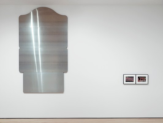 Straight-on view of the vertically-oriented, silver-metallic wall work consisting of three, horizontal rectangular panels stacked on top of one another and the two small, framed photographs on its right. Each of the panels that make up the wall work has rounded corners and a smooth surface. The top panel has a decorative curve at the top, like something one might see on a historical place marker.