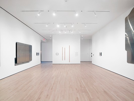 Installation view from the back wall of the gallery facing the front wall. On our left is a horizontal silver-metallic wall work with two framed photographs to its right. Directly ahead is a wall with three narrow planks of wood hung parallel to one another and a small black frame to the left of them. On our right is the wall with granite block, framed picture, and vertically-oriented silver-metallic wall work.