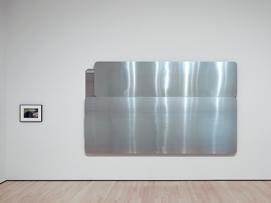 Straight-on view of the horizontal silver-metallic wall work installed on the back wall of the gallery with a small, framed photograph on its left. The silver-metallic work consists of two horizontal panels stacked on top of one another, the one on the bottom about twice the height of the one on top. The top panel has a slightly shorter width than the bottom panel, and they are both aligned on the right side, exposing a bit of another metal panel in the top left corner that is behind the front two.