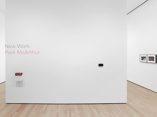 A straight-on view of a white entry wall to a museum gallery with light blonde wood floors and even lighting. The title of the exhibition, "New Work" and the artist's name, "Park McArthur" are installed on the wall in vinyl lettering, the title in grey and the artist's name in red, and both in the same thin, sans serif font. They are stacked one on top of the other and installed so that their left-most edges are flush with the edge of the wall. Beneath the title text is a box of exhibition brochures, and on the right side of the wall is a small rectangular work, its details too distant to make out.
