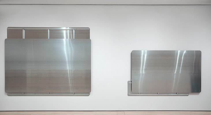Straight-on view of two silver-metallic wall works, hung so that their bottom edges are aligned horizontally on the wall. The work on the left is larger than the one on the right, and both have partially exposed back panels with structural metal beams. As with the other silver-metallic wall works, their corners are rounded and their surfaces smooth.
