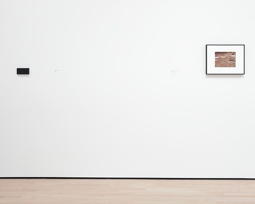 Straight-on view of two works hung on a white wall. On our left is another small, granite block, but this one does not bear any inscription. On our right is a framed photograph with a thick white mat and a thin black frame of a sandy ground.