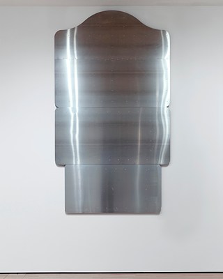 Straight-on view of the vertically-oriented, silver-metallic wall work consisting of four, horizontal rectangular panels stacked on top of one another. Each of the four panels that make up the work has rounded corners and a smooth surface. The top panel has a decorative curve at the top, like something one might see on a historical place marker.