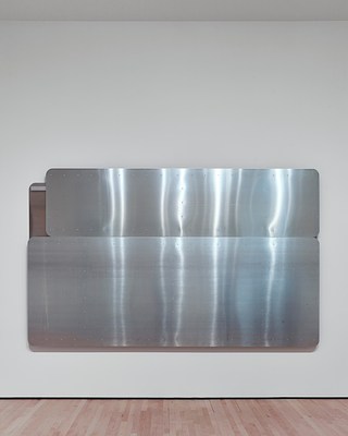 Straight-on view of the horizontal silver-metallic wall work installed on the back wall of the gallery. The work consists of two horizontal panels stacked on top of one another, the one on the bottom about twice the height of the one on top. The top panel has a slightly shorter width than the bottom panel, and they are both aligned on the right side, exposing a bit another metal panel in the top left corner that is behind the front two.
