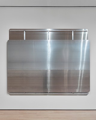 A close-up view of a large, silver-metallic wall work with partially-exposed back panel showing structural metal beams—two horizontal at top and bottom, and three vertical. At least one column of screws dotting the surface of the front panel is aligned with the vertical metal beam behind it. The surface of the sculpture reflects the overhead light in long bands.