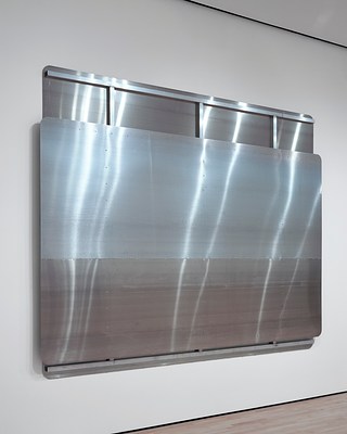 Close up left-angle view of the large, silver-metallic wall work with partially-exposed back panel showing structural metal beams—two horizontal at top and bottom, and three vertical. At least one column of screws dotting the surface of the front panel is aligned with the vertical metal beam behind it. The surface of the sculpture reflects the overhead light in long bands.