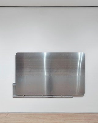 Straight-on view of the smaller of the two silver-metallic wall works hanging on this wall. Peaking out from beneath the large front panel is the back side of a smaller back panel, with parts of its structural metal beams exposed.
