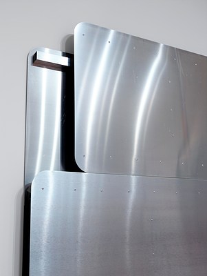 Detailed side-angle view of the silver-metallic wall work showing the top left corner where the back panel is exposed. There looks to be a depth of a few inches between front and back panels and a metal bar at the top of the back panel. Small, regularly-placed screws dot the surface of the otherwise smooth front panels.