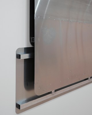 Close-up left-angle view of the bottom left corner of the silver-metallic wall work, showing the back panel sticking out from behind the front panel, and a gap of what looks like a few inches between them.