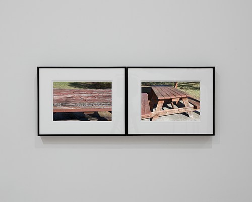 Straight-on view of two framed photographs installed side by side so that their right and left edges are touching. Both have thin, black frames and thick, white mats. The photograph on our left is a close-up view of the weather-worn surface of a faded red picnic table, a sunny patch of grass in the background. The photograph on our right shows more of the picnic table and the concrete slab on which it sits.