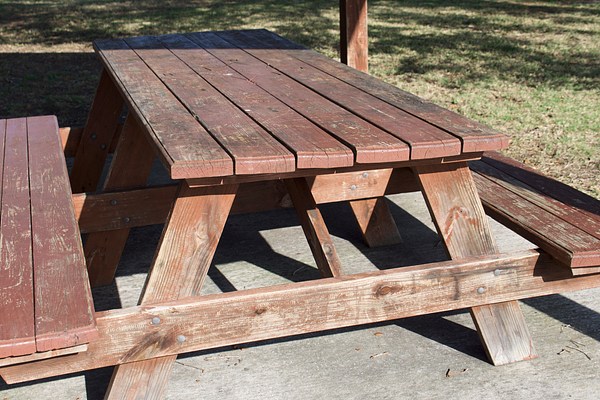 The photograph that shows a mostly-full view of the picnic table and the concrete slab on which it sits, from a skewed side angle, so that both its base and surface are visible. It is mostly in full sun and its legs cast long dark shadows on the concrete slab beneath it.