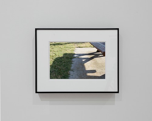 Straight-on view of a framed photograph with a thin, black frame and thick, white mat. The photograph shows the shadow cast by a picnic table on the line formed where concrete meets grass.
