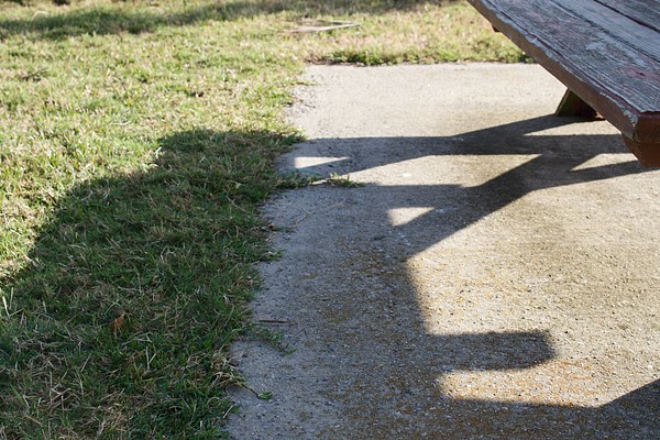 A photograph showing the shadow cast by a picnic table on the line formed where concrete meets grass.