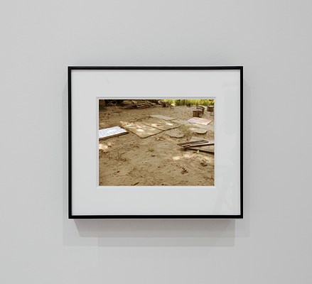 A photograph in a thin black frame with a thick white mat showing a shaded patch of sand with a bamboo mat and other wooden planks and boards strewn about on top of it. In the background is a pile of scrap wood, a semi-circle of tree stumps, and some grass lining the perimeter. A concrete sidewalk enters into the picture plane from the right side at a diagonal, ending in sand.