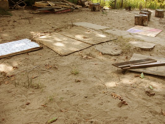 A close up of a photograph of a shaded patch of sand with a bamboo mat and other wooden planks and boards strewn about on top of it. In the background is a pile of scrap wood, a semi-circle of tree stumps, and some grass lining the perimeter. A concrete sidewalk enters into the picture plane from the right side at a diagonal, ending in sand.